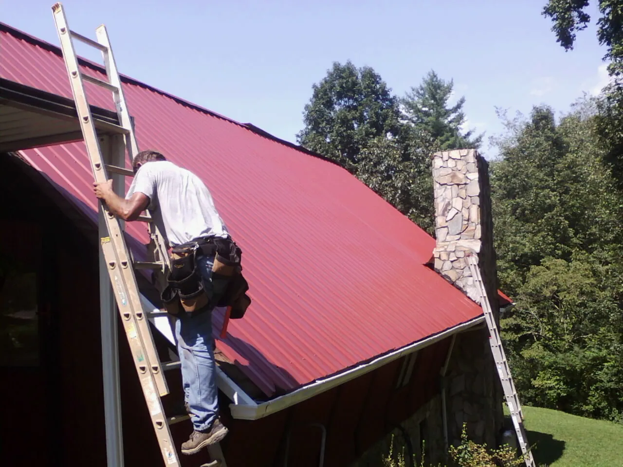 Roofer on a ladder beside a red metal roof and stone chimney — Soffit & Fascia Repair by Roofing Brothers in Stockbridge